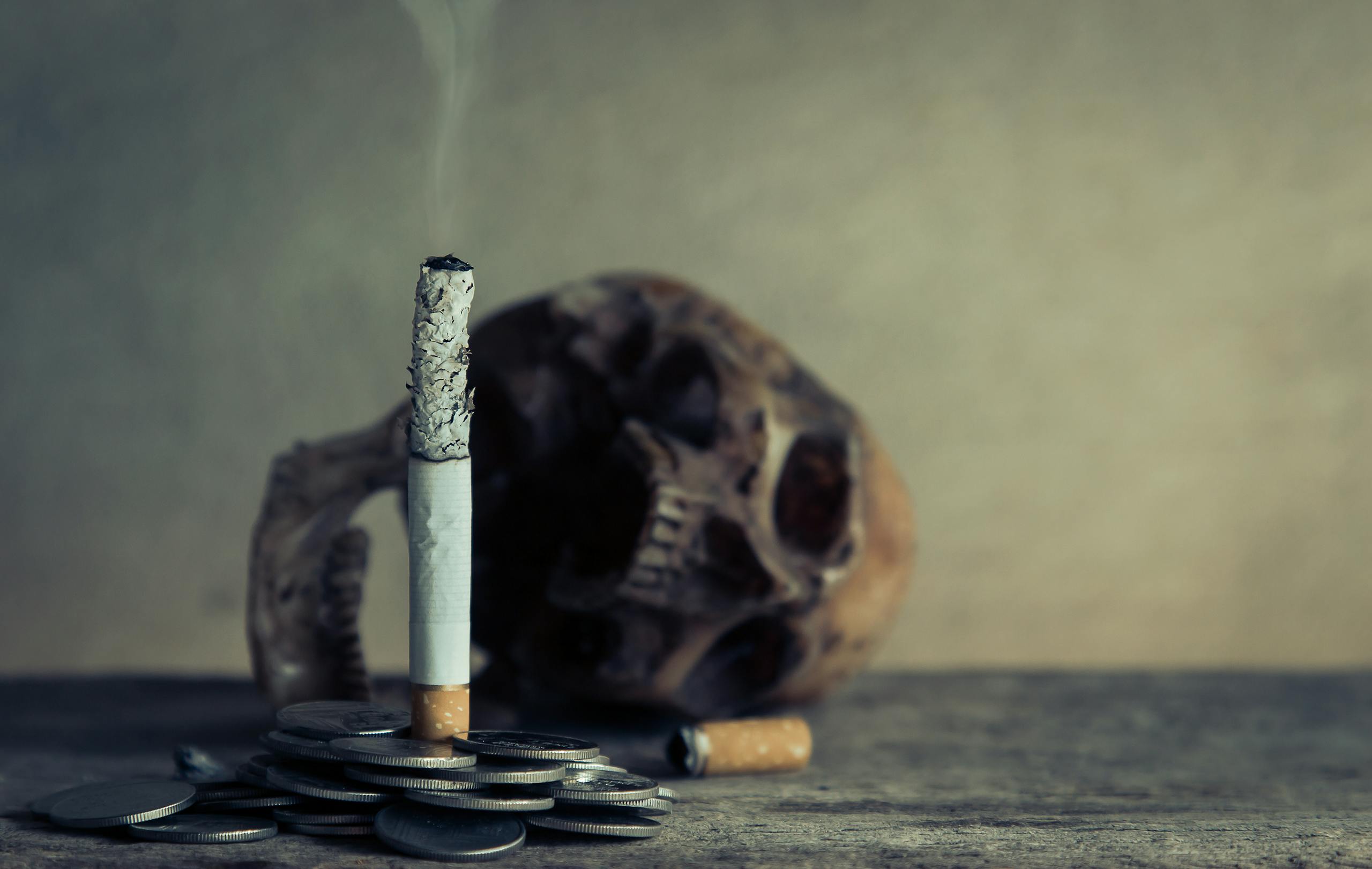 A cigarette burning on coins next to a skull, illustrating the concept of smoking and death.