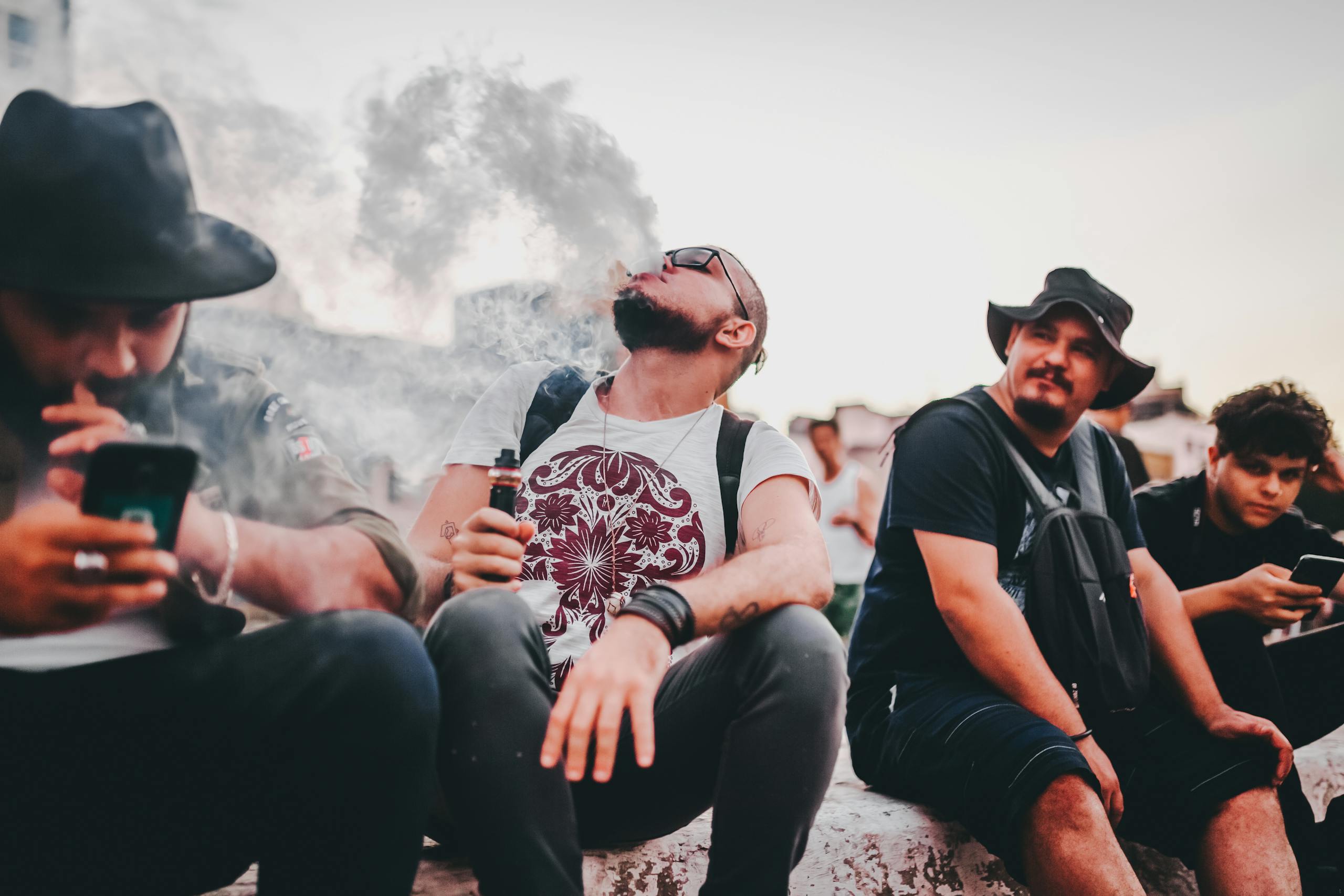 A group of men sitting outdoors, vaping and using mobile phones, in a casual setting.