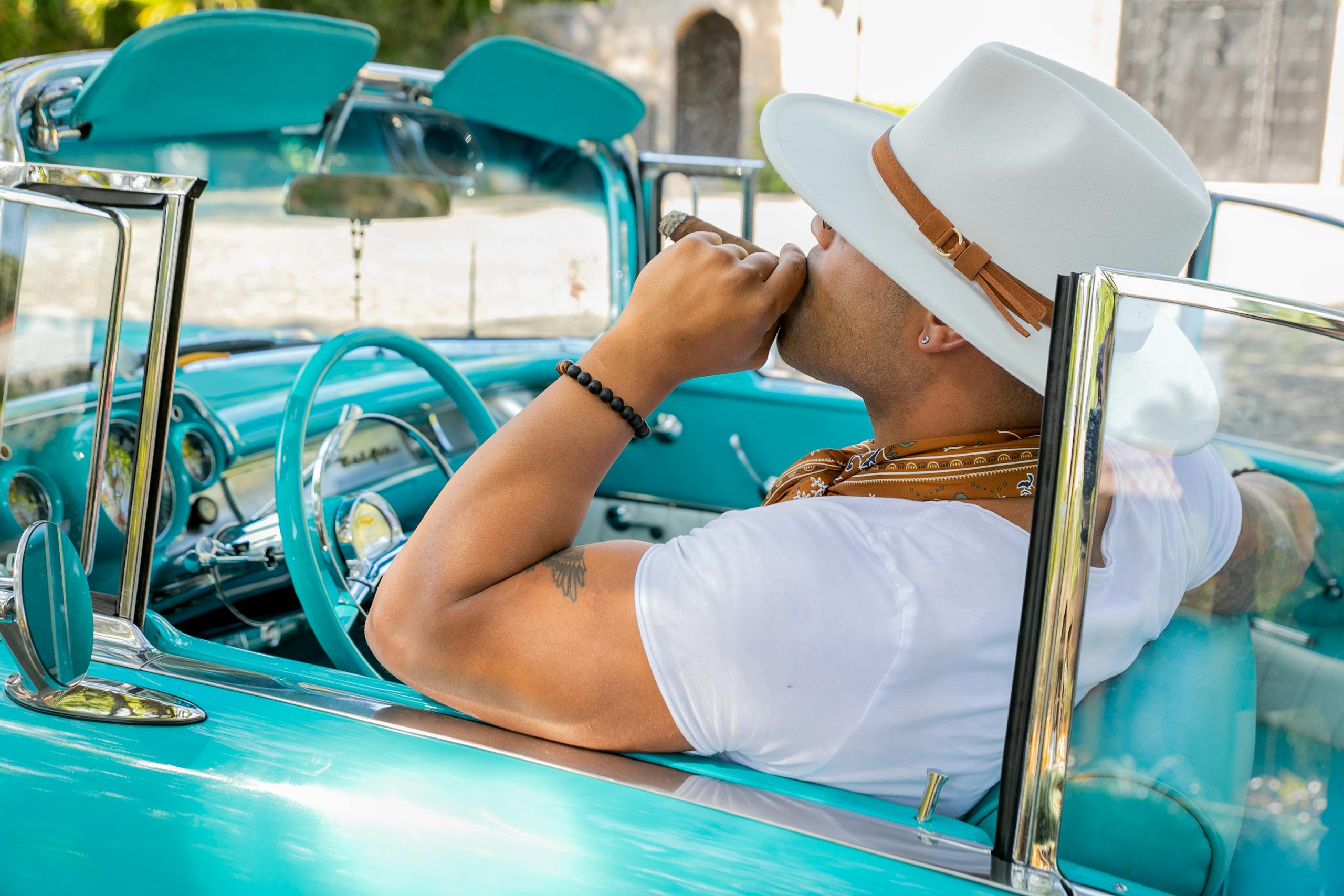 A man enjoying leisure with a cigar, wearing a hat, in a vintage convertible car.