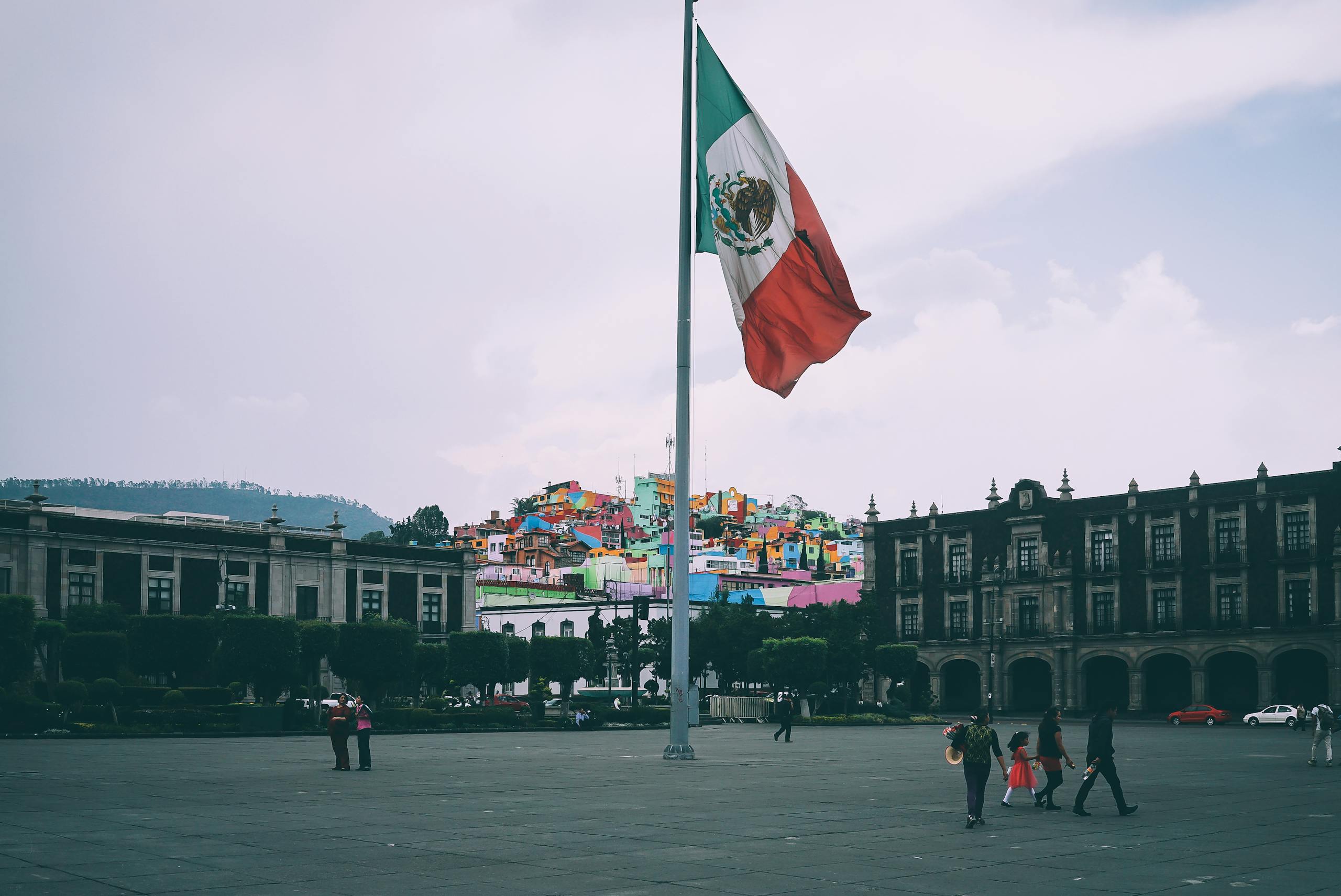 Colorful cityscape of Toluca with large Mexican flag in the foreground, vibrant buildings in the backdrop.