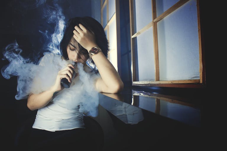 Cool casual brunette in white t shirt smoking electronic cigarette while leaning on hand in dark studio