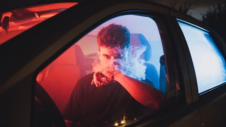 Moody image of a young man vaping inside a car with vibrant red and blue lighting.