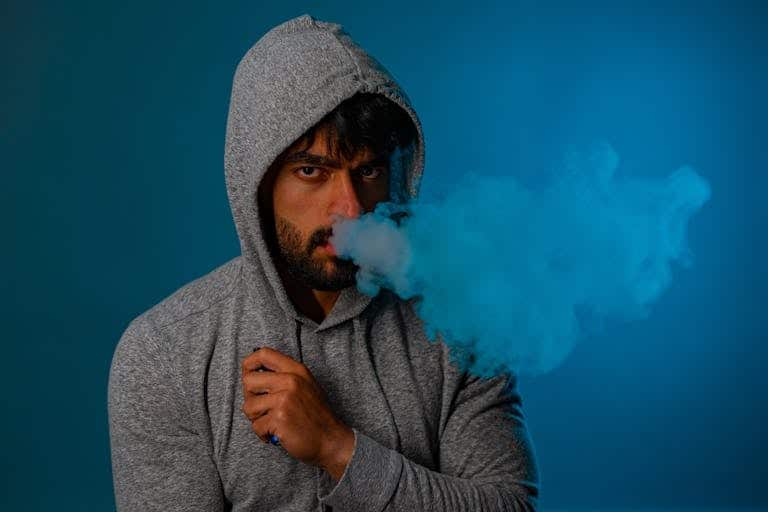 Portrait of a man in a grey hoodie exhaling blue vape smoke against a studio background.