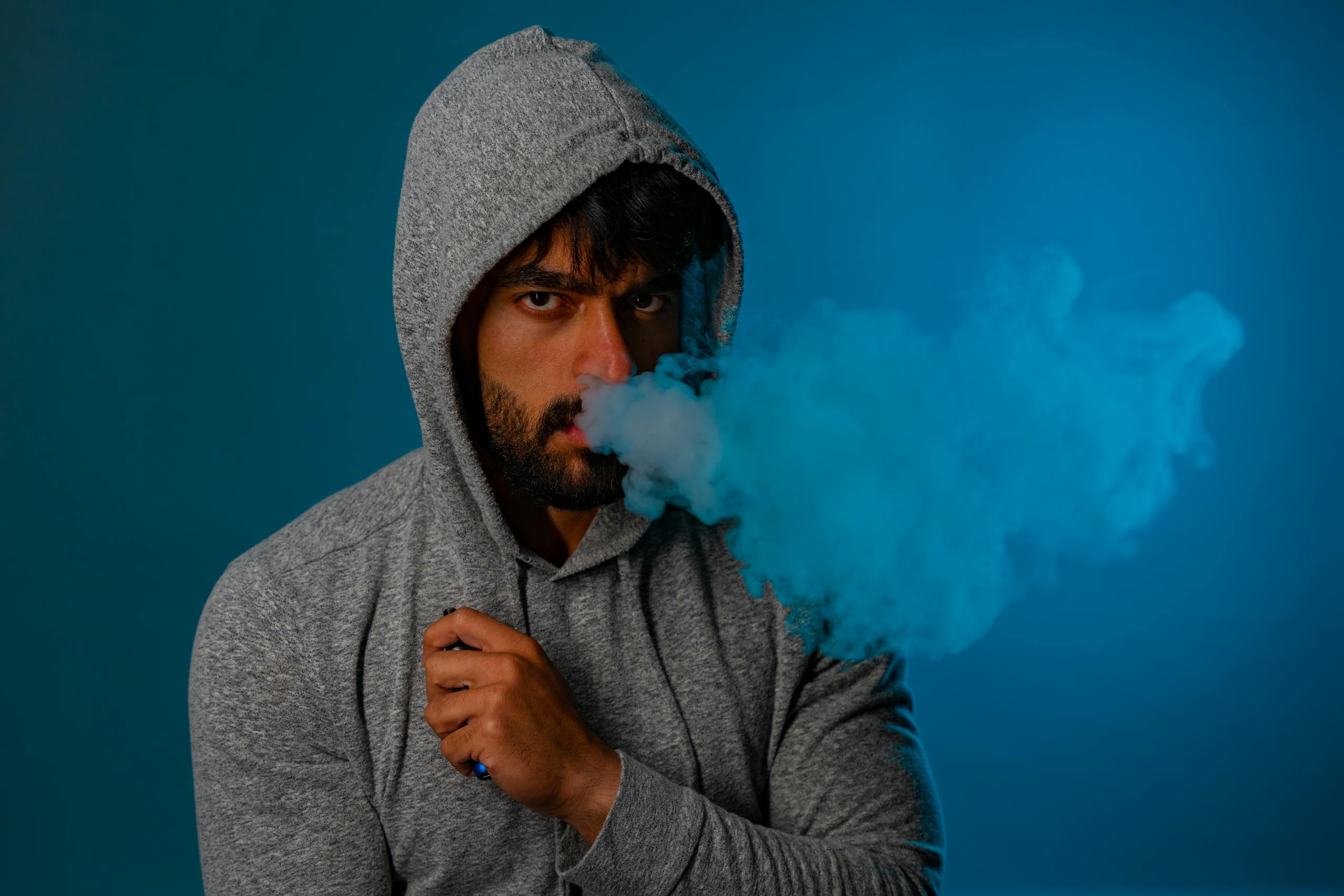 Portrait of a man in a grey hoodie exhaling blue vape smoke against a studio background.