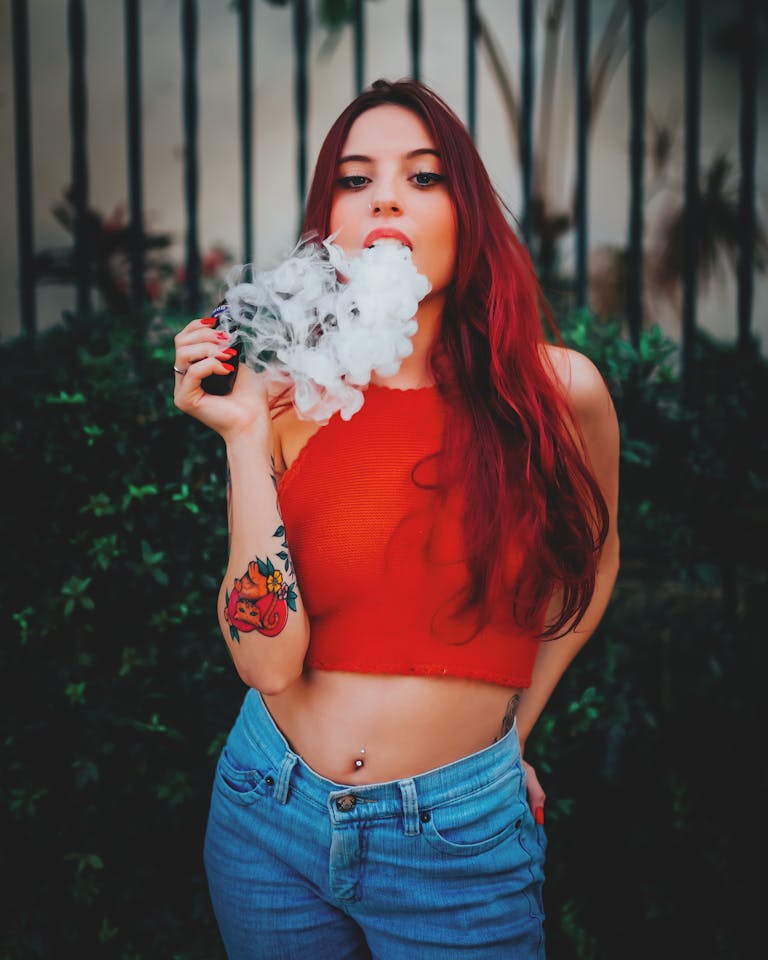 Young woman with red hair vaping outdoors, wearing a red top and jeans, showcasing tattoos.